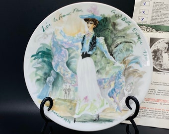 Pode incluir: Um prato de porcelana branca com uma pintura em aquarela de uma mulher com um vestido azul e um lenço de estampa floral. O prato é intitulado "Lea la femme-Fleur" e "La Belle Epoque 1900" e é assinado "Ganev 1976".