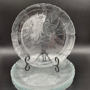 May include: A set of four clear glass plates with a delicate floral design. The plates are stacked on top of each other, with the top plate featuring a more prominent floral pattern.
