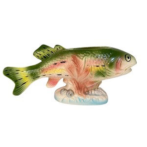 Puede incluir: Figura de pez de cerámica en tonos verdes, amarillos, rosas y azules. El pez tiene escamas y aletas detalladas, con una pequeña abertura en la parte superior. La figura descansa sobre una base con un diseño de olas.