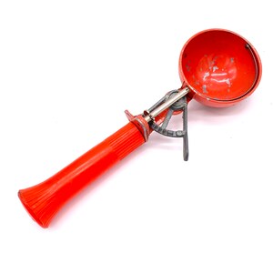 Op de afbeelding: Een rode metalen ijslepel met een lange oranje kunststofsteel. De lepel heeft een ronde kom en een metalen hendel aan de zijkant.