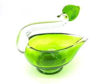 May include: A green glass decorative bowl with a clear handle and a green leaf accent. The bowl has a pedestal base and a curved handle. The leaf is a bright green color.