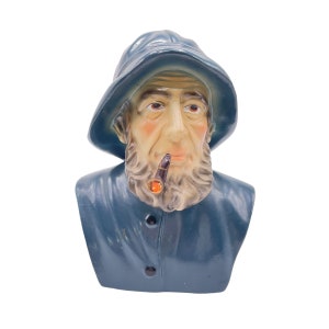 May include: A blue ceramic bust of a man wearing a blue hooded jacket and a pipe. The man has a white beard and mustache and is looking straight ahead.