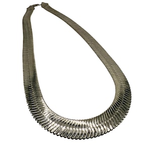 Puede incluir: Un collar plateado con un diseño plano de espiga. El collar está hecho de piezas metálicas entrelazadas, creando una superficie flexible y texturizada. El cierre es visible en la parte superior y el collar se curva con gracia.