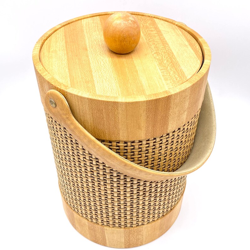 May include: A wooden ice bucket with a woven wicker design and a wooden lid with a knob. The bucket has a brown leather handle.