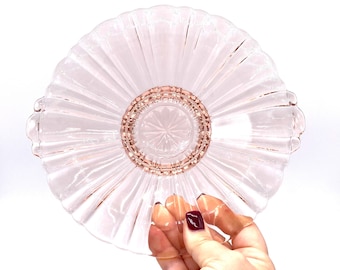 Puede incluir: Un plato de servir de vidrio transparente con un borde rosa y un borde festoneado. El plato tiene un diseño acanalado y un centro elevado.