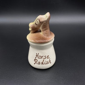 May include: A ceramic horse radish jar with a horse head lid. The jar is off-white with brown text that reads "Horse Radish". The lid is brown and tan. The jar is on a black surface.