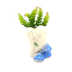 Könnte beinhalten: Eine herzförmige Keramikvase in Creme mit blauer Schleife und goldenen Verzierungen, die eine grüne Sukkulentenpflanze enthält. Die Vase hat ein Blumendesign. Die Gesamtästhetik ist vintage und dekorativ.
