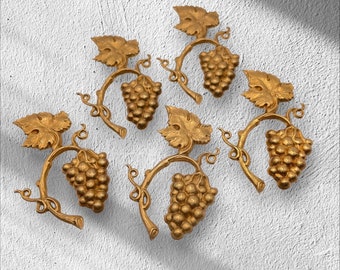 Könnte beinhalten: Sechs goldfarbene Wanddekorationen in Form von Weinreben. Jedes Stück zeigt eine Traube, Blätter und einen gewundenen Stiel. Die Dekorationen sind an einer strukturierten weißen Wand angebracht und erzeugen einen klassischen, eleganten Look.