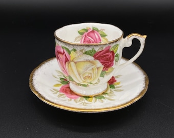 May include: A white teacup and saucer set with a floral design featuring pink, red, and yellow roses. The set has a gold trim around the edges.