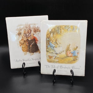 May include: Two framed illustrations of Beatrix Potter's children's books. The first illustration shows two rabbits walking in a snowy landscape. The second illustration shows a rabbit family sitting in a garden. The text on the frames reads "Appley Dapply's Nursery Rhymes" and "The Tale of Benjamin Bunny".