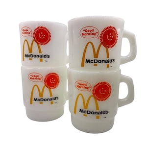 Op de afbeelding: Set van vier witte keramische mokken met een gele McDonald's-boog en een rode zon met een smiley. De mokken hebben de tekst "Good Morning" in een spreekballon.