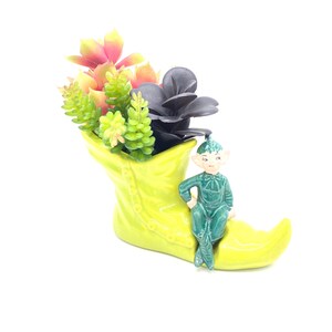 Può includere: Un vaso da fiori a forma di stivale verde in ceramica con una piccola statuina di elfo verde seduta sul bordo. Il vaso è pieno di una varietà di piante grasse, tra cui verdi, rosa e nere.