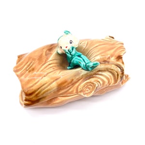 Può includere: Un piatto in ceramica a forma di tronco d'albero con una statuetta di fata verde e bianca seduta sopra. La fata indossa un vestito verde e ali bianche.