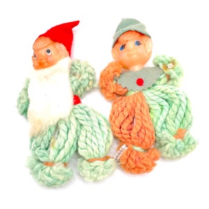 Puede incluir: Dos adornos de gnomo de lana hechos a mano. Un gnomo tiene un gorro rojo y lana verde, el otro tiene un gorro verde y lana naranja y verde.