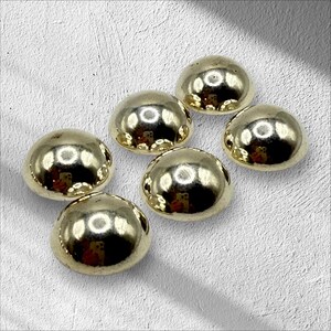 May include: Six gold-colored, dome-shaped buttons are arranged on a textured, off-white surface. The buttons have a shiny, reflective finish, and their rounded shape is prominent. The buttons are likely for sewing or crafting projects.