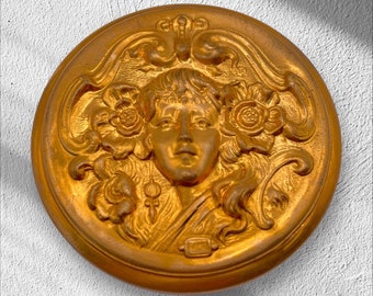 May include: A round, antique-style, gold-colored decorative button with a detailed relief of a woman's face surrounded by floral and ornate scrollwork. The button is set against a textured white background.