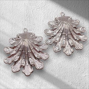 May include: Two silver-toned metal shell-shaped charms with a detailed, textured design. The charms have a loop at the top for attaching to jewelry or other crafts.