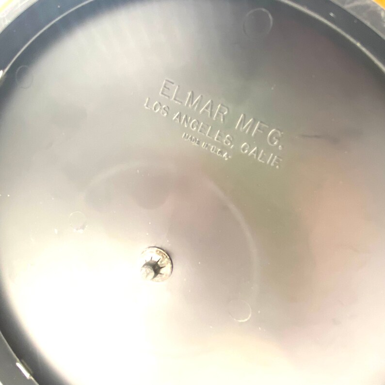 May include: Close-up of the bottom of a gray plastic object with a silver screw. The object has a black label that reads "ELMAR MFG. LOS ANGELES, CALIF. MADE IN U.S.A."