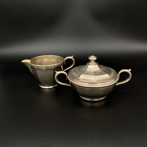 May include: A silver creamer and sugar bowl set. The creamer has a fluted design and a handle. The sugar bowl has a lid and a rounded shape.
