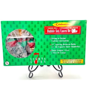Könnte beinhalten: Eine grüne Schachtel mit Celebrations-Blasenlichtern. Die Schachtel hat einen grünen Hintergrund mit kleinen weißen Weihnachtsbäumen. Die Schachtel enthält sieben Blasenlichter mit roten und grünen Glühbirnen. Die Gesamtlänge beträgt 2,13 Meter.