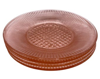 Puede incluir: Una pila de cuatro platos de cristal rosa. Cada plato tiene un borde exterior acanalado y un centro texturizado con forma de diamante. Los platos están apilados uno encima del otro, mostrando su diseño.