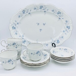 May include: A white porcelain dinnerware set with a blue floral pattern and silver trim. The set includes a large oval platter, teacups, saucers, and small bowls. The teacups have silver rims and handles.