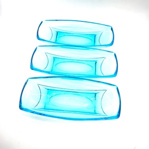 Puede incluir: Tres platos de servir de vidrio azul apilados uno encima del otro. Los platos son rectangulares con bordes redondeados y una ligera curva en el medio.