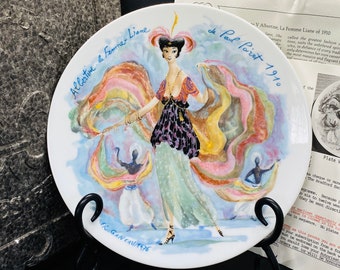 May include: A white porcelain plate with a colorful illustration of a woman in a flowing dress and a feathered hat. The plate is titled "Albertine & Femme-Liane" and "de Paul Poiret 1910".