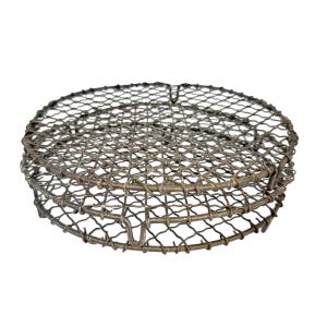 May include: A round, metal wire mesh cooking grate. The grate is made of interwoven metal wires, forming a tight mesh pattern. It has a circular shape with a slightly raised edge and is designed for grilling or cooking.