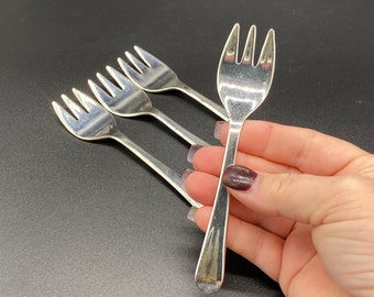 May include: Four silver dessert forks with a polished finish. The forks are held in a hand, with one fork in the foreground.
