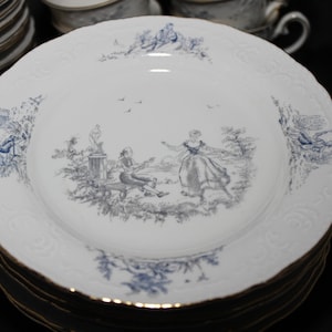 Può includere: Un set di piatti piani in porcellana bianca con un motivo floreale blu e grigio. I piatti presentano una scena di una coppia in un giardino.