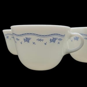 May include: Set of four white ceramic mugs with a blue floral pattern around the rim. The mugs have a simple, classic design.