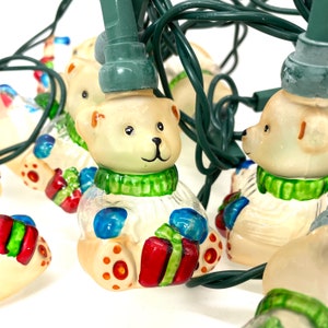 May include: A string of Christmas lights featuring white ceramic bear ornaments with green, red, and blue accents. Each bear is holding a gift box.