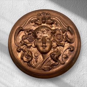 Puede incluir: Una escultura en relieve circular de color bronce que representa el rostro de una mujer rodeado de flores y adornos decorativos. La mujer tiene el pelo largo y adornos florales. Una obra de arte detallada de estilo antiguo.