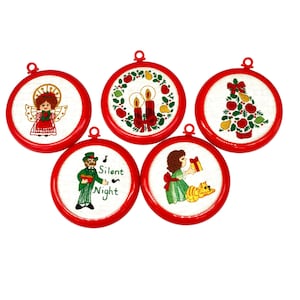 Puede incluir: Conjunto de cinco adornos navideños bordados con marco rojo. Los adornos presentan una variedad de temas navideños, incluyendo un árbol de Navidad, una corona con velas, un ángel, un cantante de villancicos y una niña con un gato.
