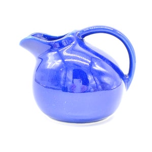 May include: A cobalt blue ceramic jug with a rounded body and a curved handle. The jug has a glossy finish.