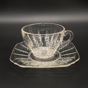 May include: A clear glass teacup and saucer set with a delicate, intricate pattern. The cup has a rounded shape and a handle, while the saucer has a scalloped edge.