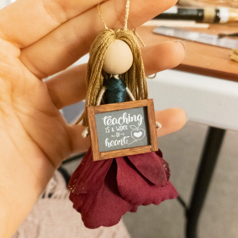 May include: A handmade doll ornament with long blonde hair, a green dress, and a burgundy flower skirt. The doll is holding a small chalkboard sign that says "Teaching is a work of heart" with a red apple graphic.