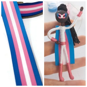 May include: A handmade doll with a white body, black hair, and a blue and pink transgender pride flag cape. The doll is wearing a blue and pink mask and has brown yarn arms and legs.