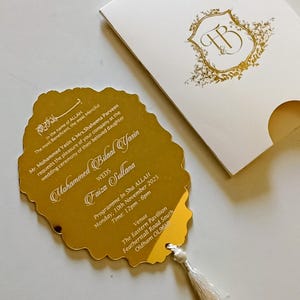 May include: A gold-colored wedding invitation with a decorative tassel, featuring event details and names. The invitation is paired with a white envelope with a gold floral crest and initials. The invitation is for a wedding ceremony.