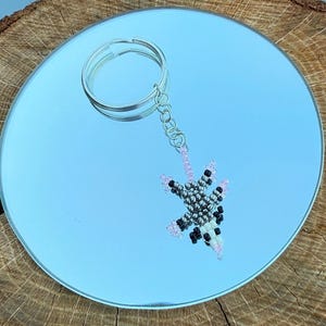 May include: A beaded keychain featuring a possum design. The possum is crafted with silver, black, white, and pink beads. The keychain has a silver ring and chain for attaching to keys or bags.