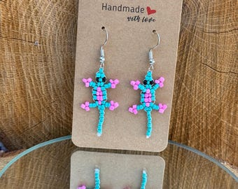 May include: Turquoise and pink beaded lizard earrings, displayed on a brown card with the text "Handmade with love". The earrings have silver-coloured hooks. The earrings are handmade.