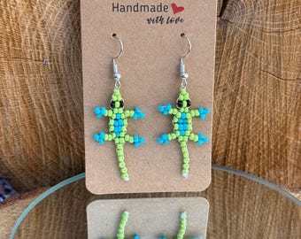 May include: Handmade beaded lizard earrings in green, blue, and black. The earrings are displayed on a brown card with the text "Handmade with love". The earrings are reflected in a mirror.