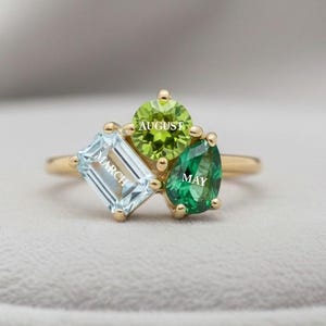 May include: A gold ring with three gemstones: a light blue emerald-cut stone with "MARCH" inscribed, a round green stone with "AUGUST", and a green pear-shaped stone with "MAY". The ring is set against a soft, neutral background.
