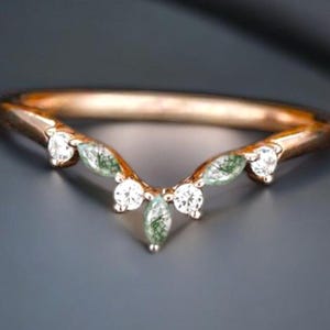 Peut inclure: Une bague en or rose avec un design incurvé. La bague présente des pierres précieuses rondes et transparentes et des pierres vertes en forme de feuille. La bague est posée sur un fond gris foncé.