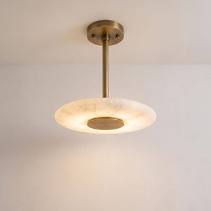 May include: A ceiling-mounted light fixture featuring a circular, translucent white stone shade with subtle veining. The fixture includes a central brass-toned metal stem and a matching circular canopy, with a small brass disc at the center of the shade.