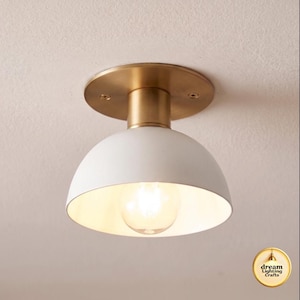 Può includere: Una plafoniera a incasso con paralume a cupola bianco opaco e base circolare in ottone spazzolato. L'apparecchio include una singola lampadina a vista e un piccolo logo circolare nell'angolo con la scritta dream Lighting Crafts.