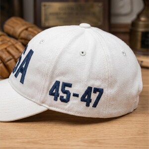 以下が含まれることがあります： 「USA」の文字と「45-47」の数字がネイビーブルーで刺繍された白い野球帽。帽子はつばがカーブしており、上部にボタンが付いています。帽子は質感のある生地で作られています。