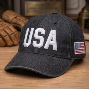 以下が含まれることがあります： 白のブロック体で「USA」の文字が入った濃いグレーの野球帽。帽子の側面には小さなアメリカ国旗のパッチが付いています。帽子は質感のある生地で作られており、つばがカーブしています。