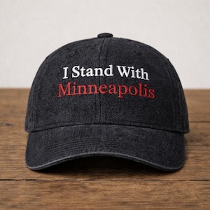 May include: A dark grey denim baseball cap featuring the embroidered phrase "I Stand With Minneapolis" in white and red. The cap has a curved peak and a button on top. The background is a wooden surface.